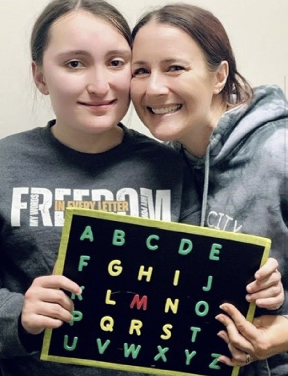 A young speller in a 'Freedom in every letter' shirt holding a colorful alphabet letterboard, cheek-to-cheek with her smiling mother.